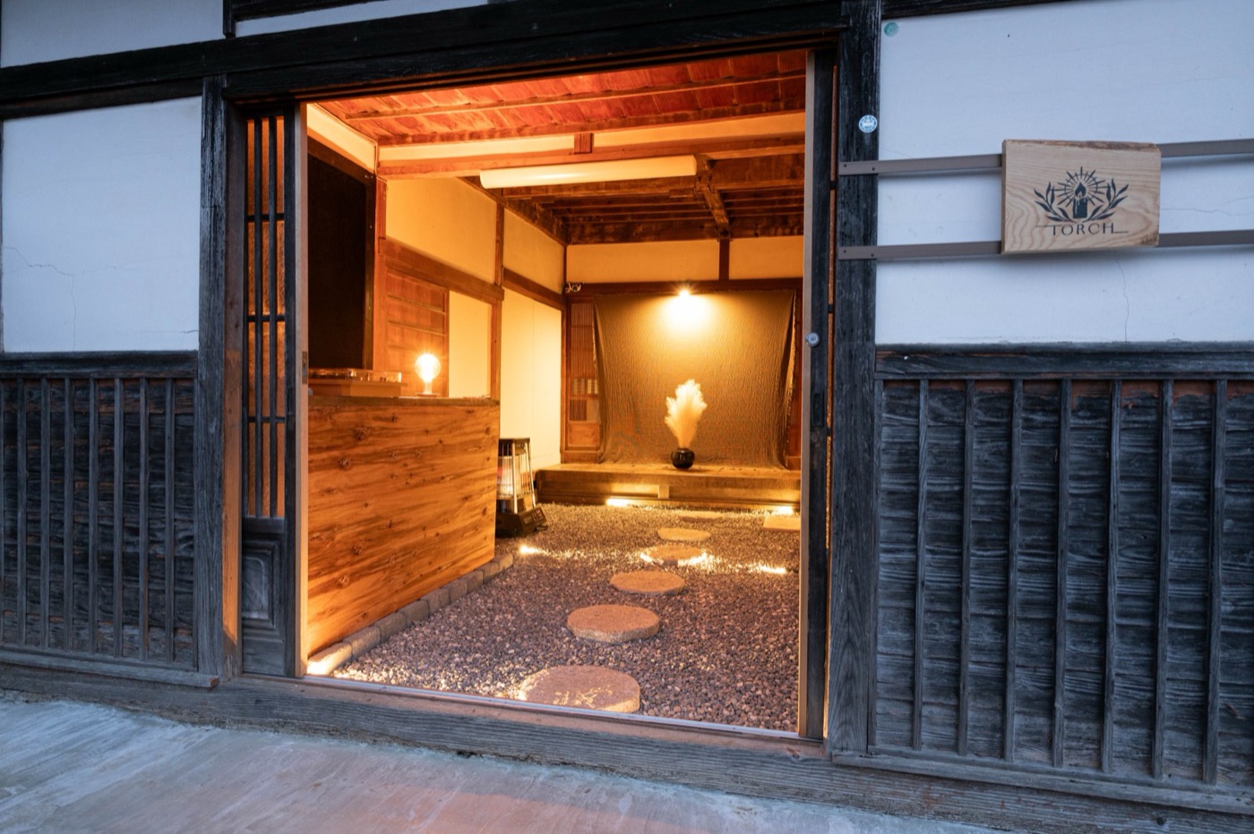 宿の玄関外観。板壁に「TORCH」の看板が掛かり、内の灯りがこぼれる